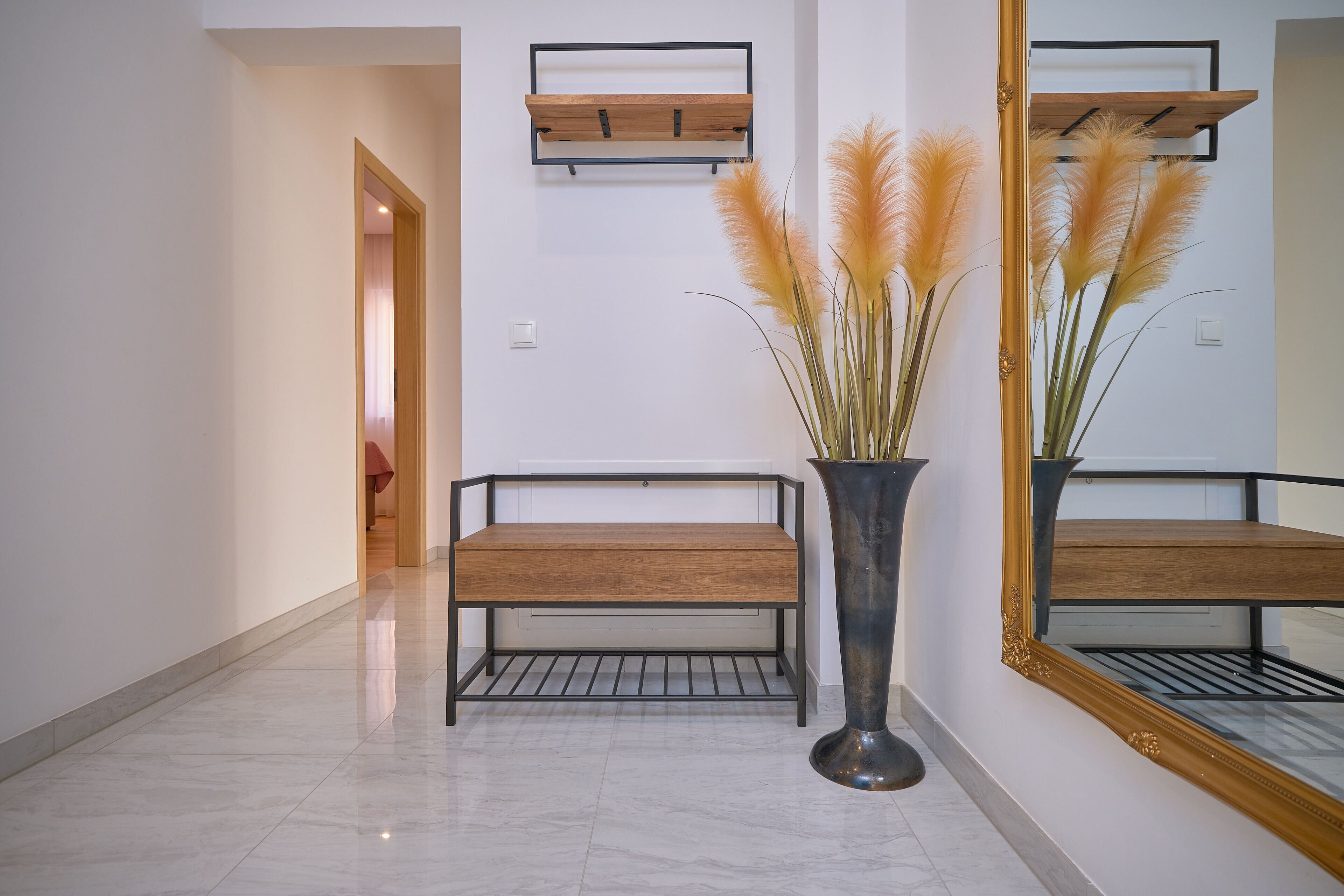 A hallway with a bench, a tall vase with pampas grass, and a large mirror on the wall.