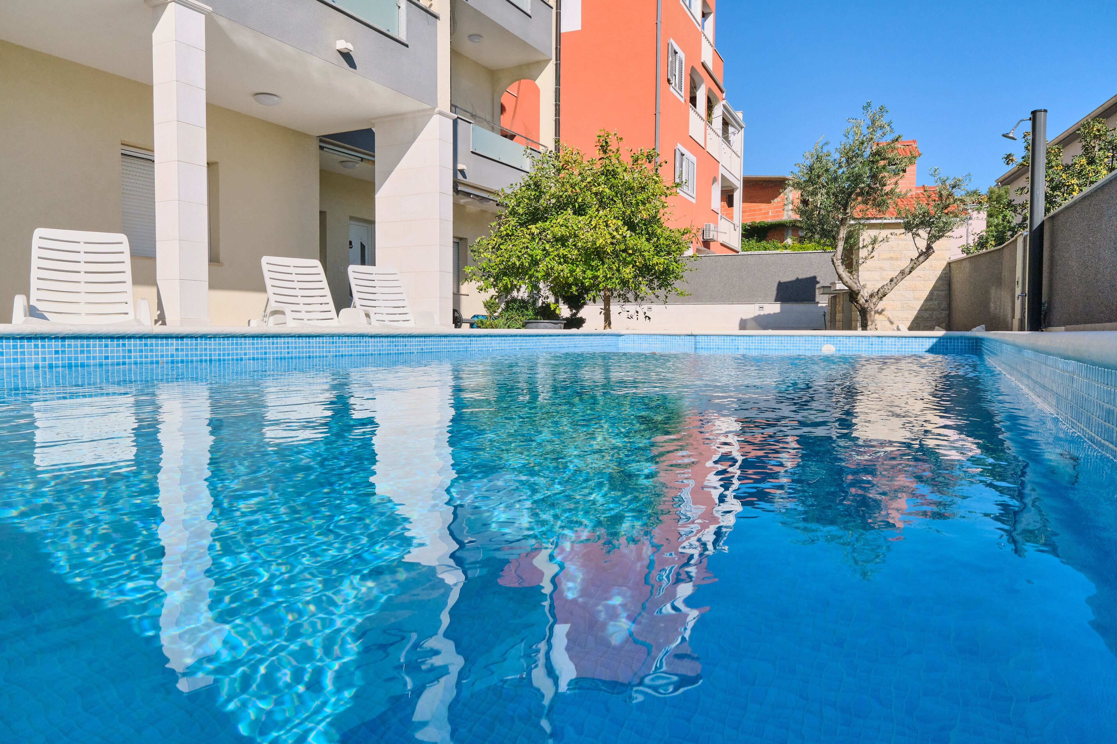 A swimming pool with clear blue water, surrounded by a building with balconies and a tree.