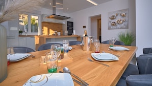 A modern dining area with a wooden table set for a meal, a kitchen in the background, and a view of the outside.