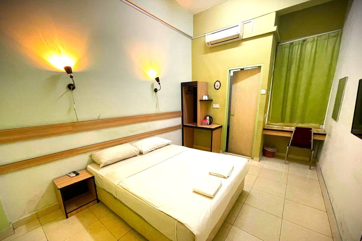 A hotel room with a bed, bedside table, lamp, desk, chair, and an air conditioning unit.