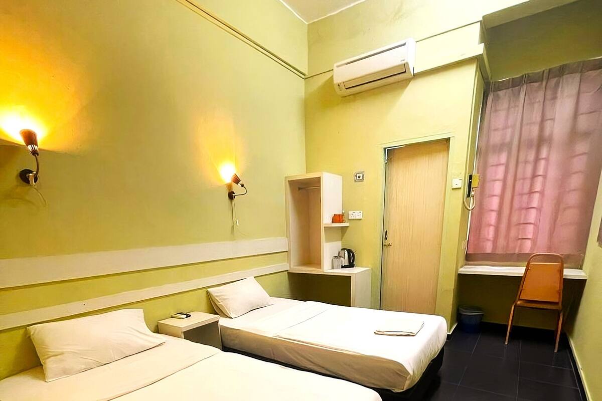 A hotel room with two beds, a small desk, a chair, an air conditioning unit, and a door with a curtain.