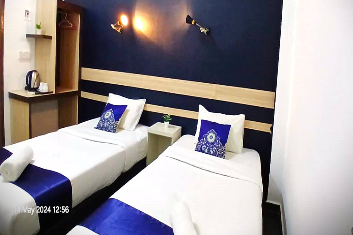 A hotel room with two single beds, a nightstand, a lamp, and a shelf with a kettle and a plant.