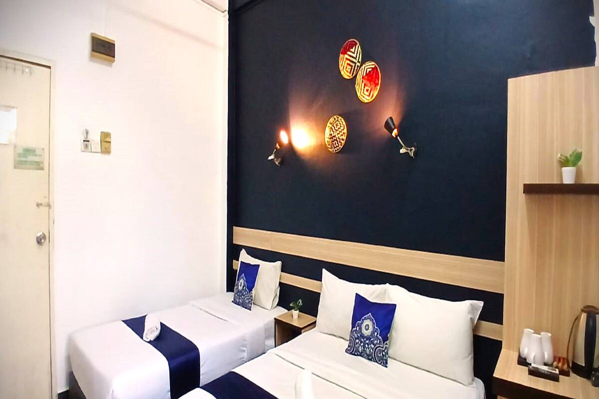 A hotel room with two beds, a dark wall with decorative wall lights, a wooden shelf with a plant, and a coffee machine.