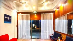 Un vestíbulo de hotel con un sofá rojo, un sofá azul y una mesa de madera con una planta. Hay cortinas en las ventanas y un cartel en la pared.