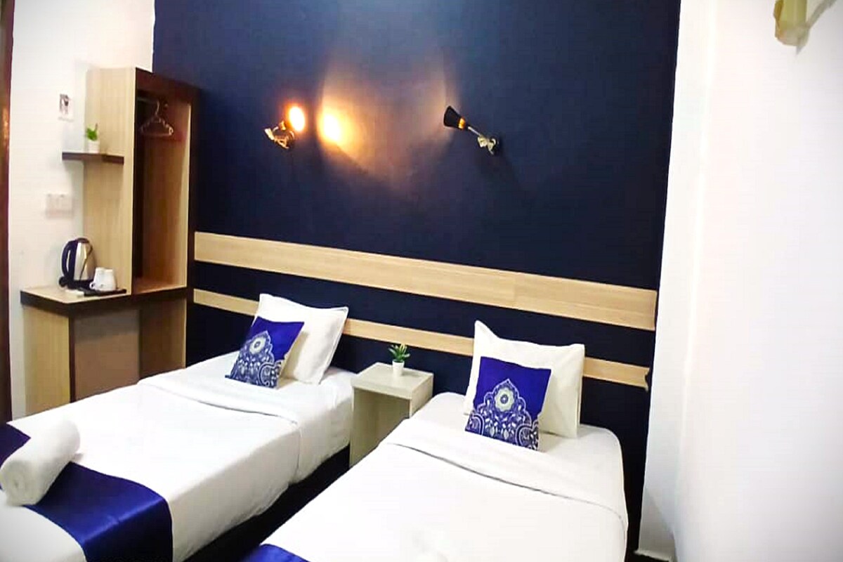 A hotel room with two single beds, a nightstand, a lamp, and a shelf with a kettle and a plant.