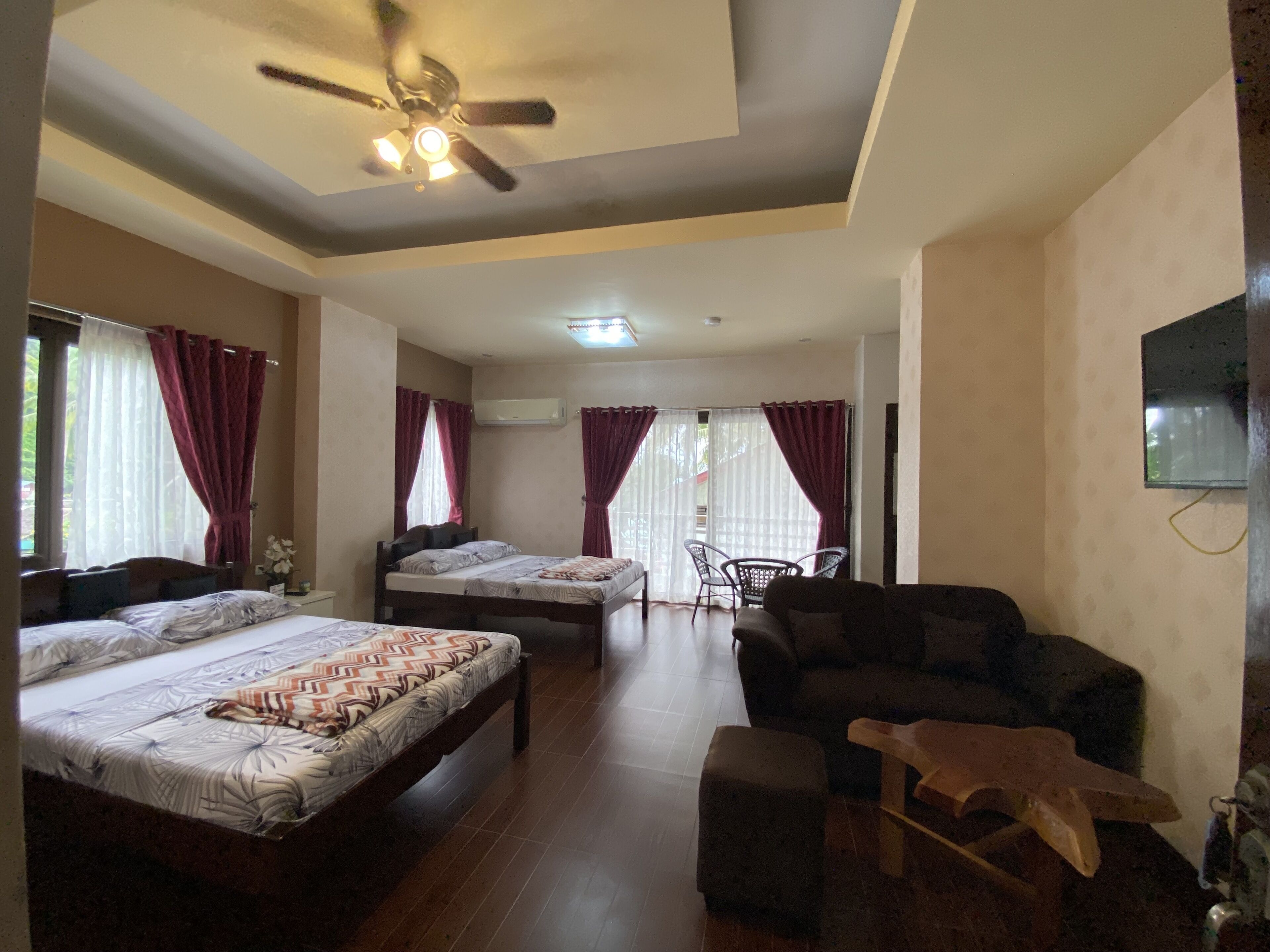 Habitación de hotel con dos camas, ventilador de techo, sofá y televisor montado en la pared.