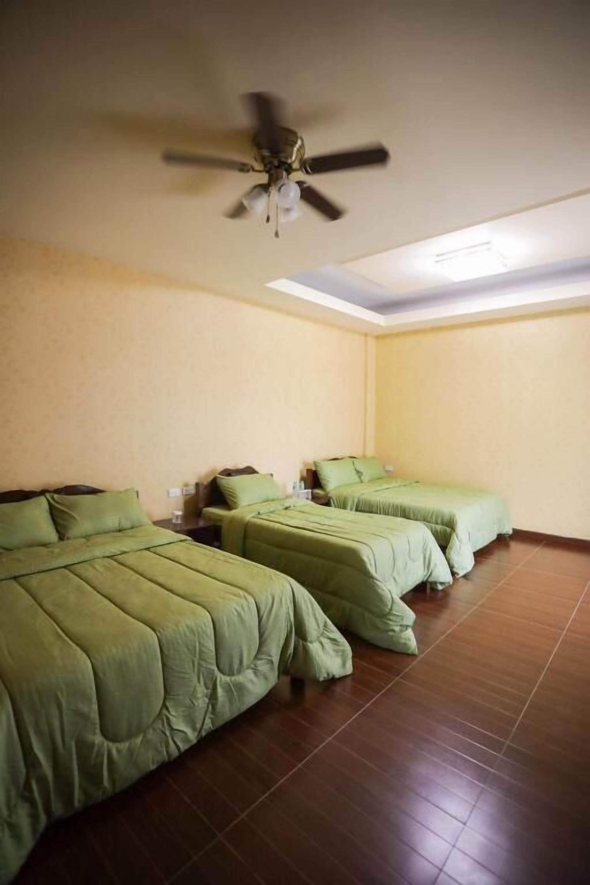 A room with three beds, each with green bedding, a ceiling fan, and a tiled floor.