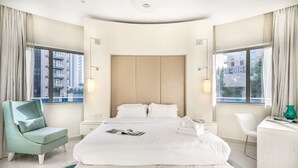 Una habitación de hotel moderna con una cama grande, una silla, un escritorio y vistas de la ciudad a través de amplias ventanas.
