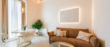 A modern living room with a brown sofa, a round glass coffee table, a white dining table with chairs, and a potted plant.