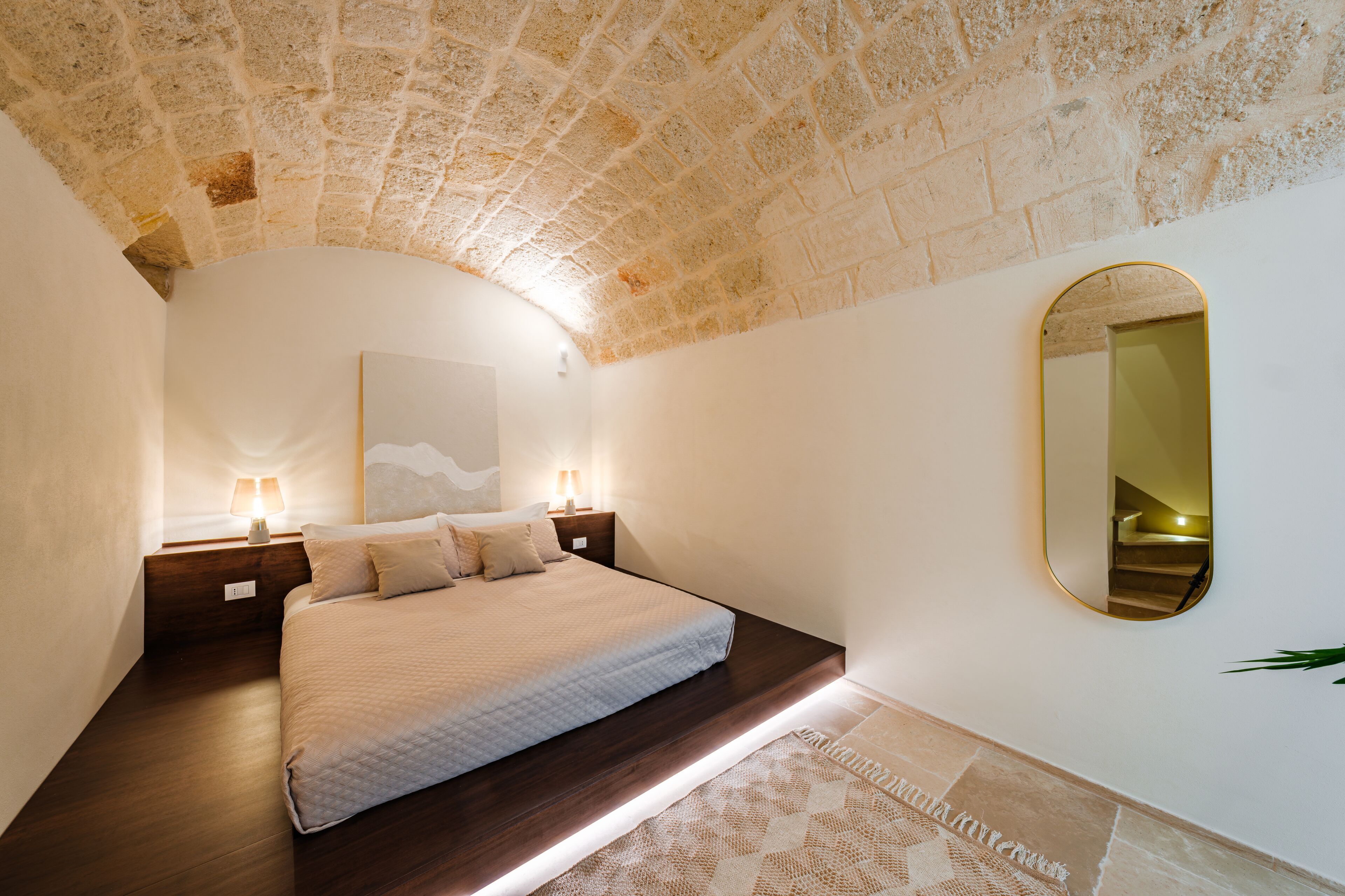 A bedroom with a stone ceiling, a bed with a headboard, bedside lamps, a large mirror, and a patterned rug.