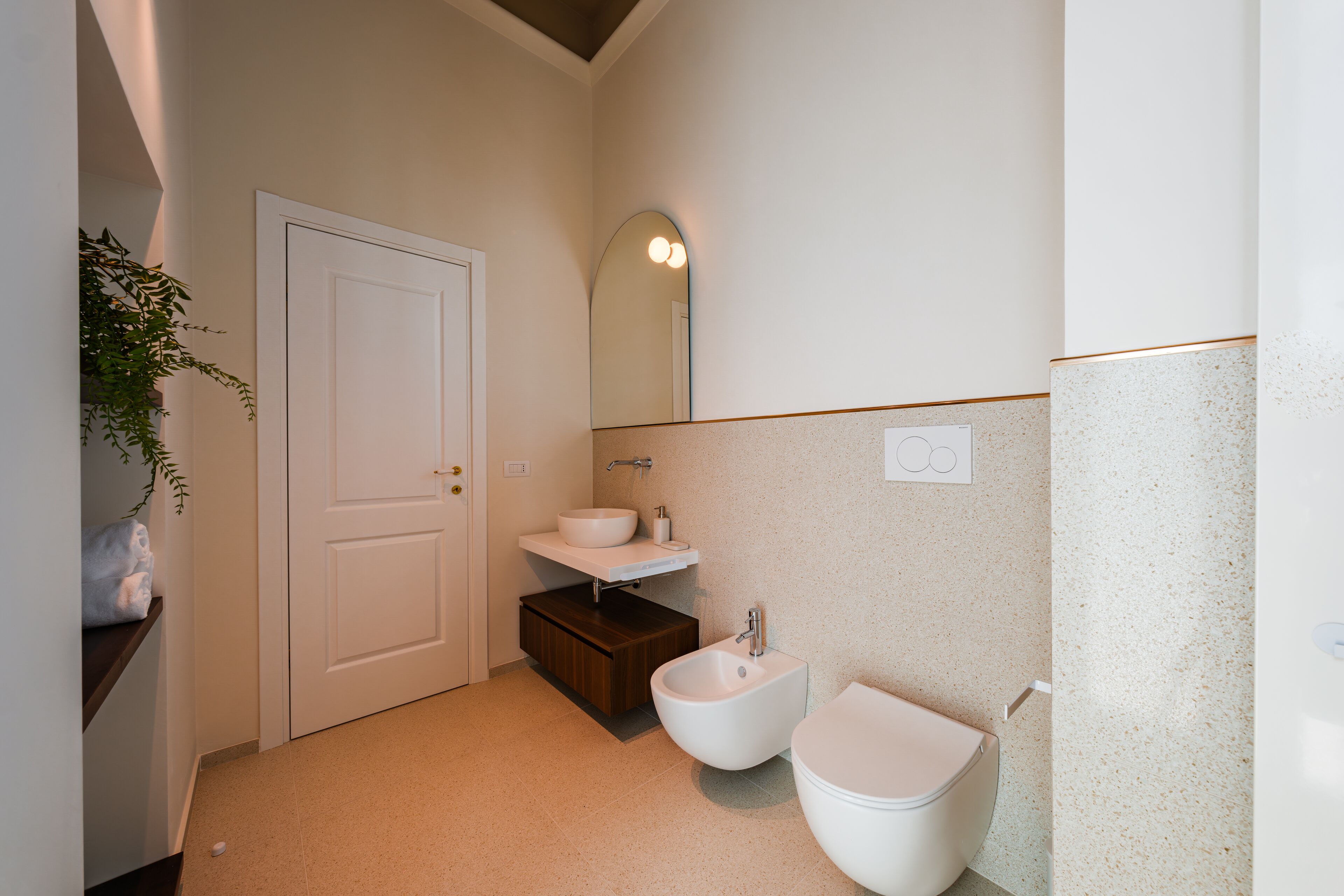 A bathroom with a white toilet, a bidet, a sink with a mirror above it, and a plant on the left.