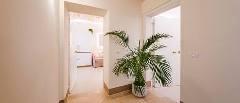 A hallway with a potted palm plant, a light fixture on the wall, and a door leading to a room with a bed and a plant.