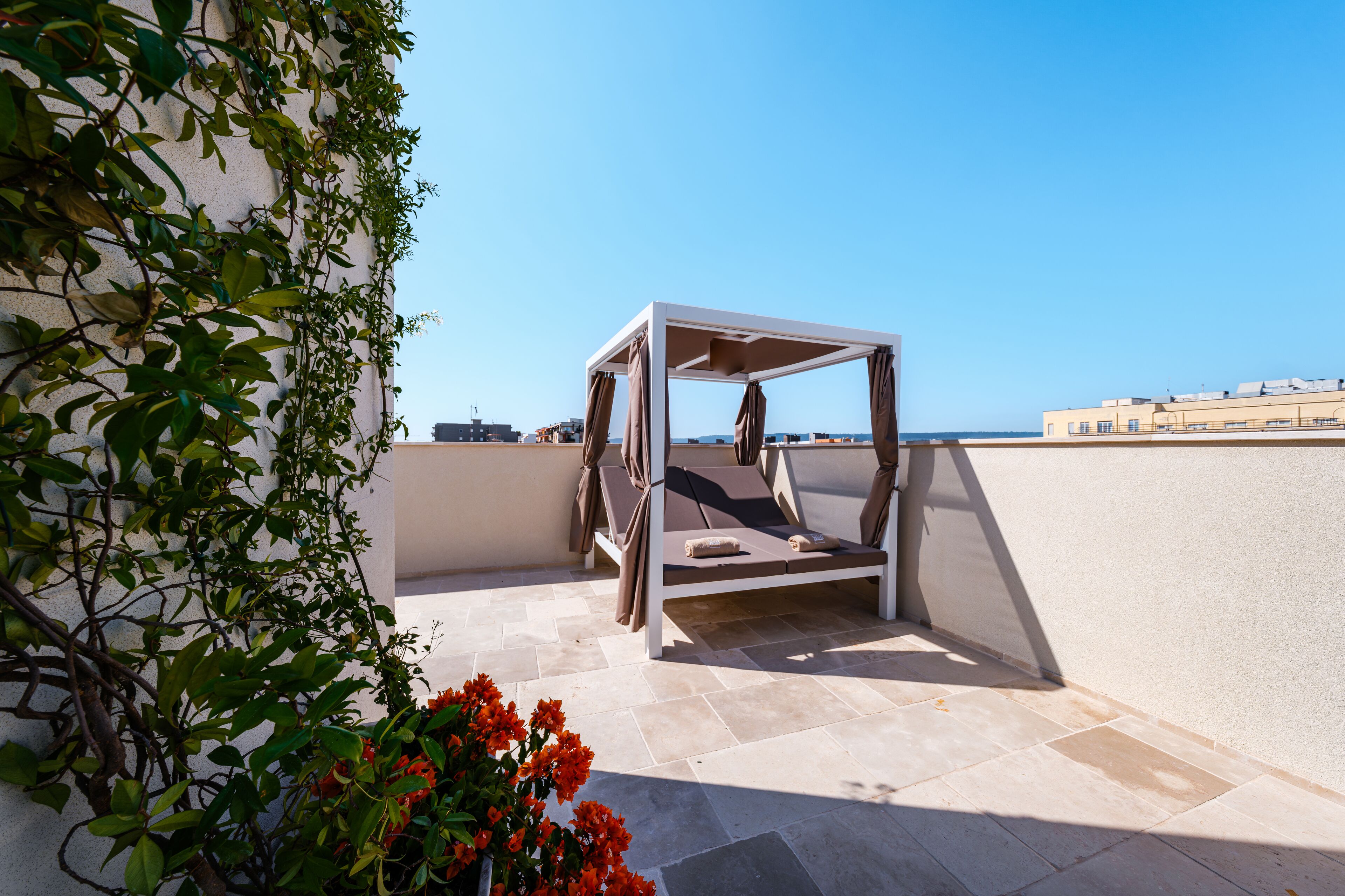 A sunlit outdoor seating area with a white metal structure, beige tiled flooring, and a view of the sea and buildings in the background.