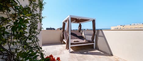 A sunlit outdoor seating area with a white metal structure, beige tiled flooring, and a view of the sea and buildings in the background.