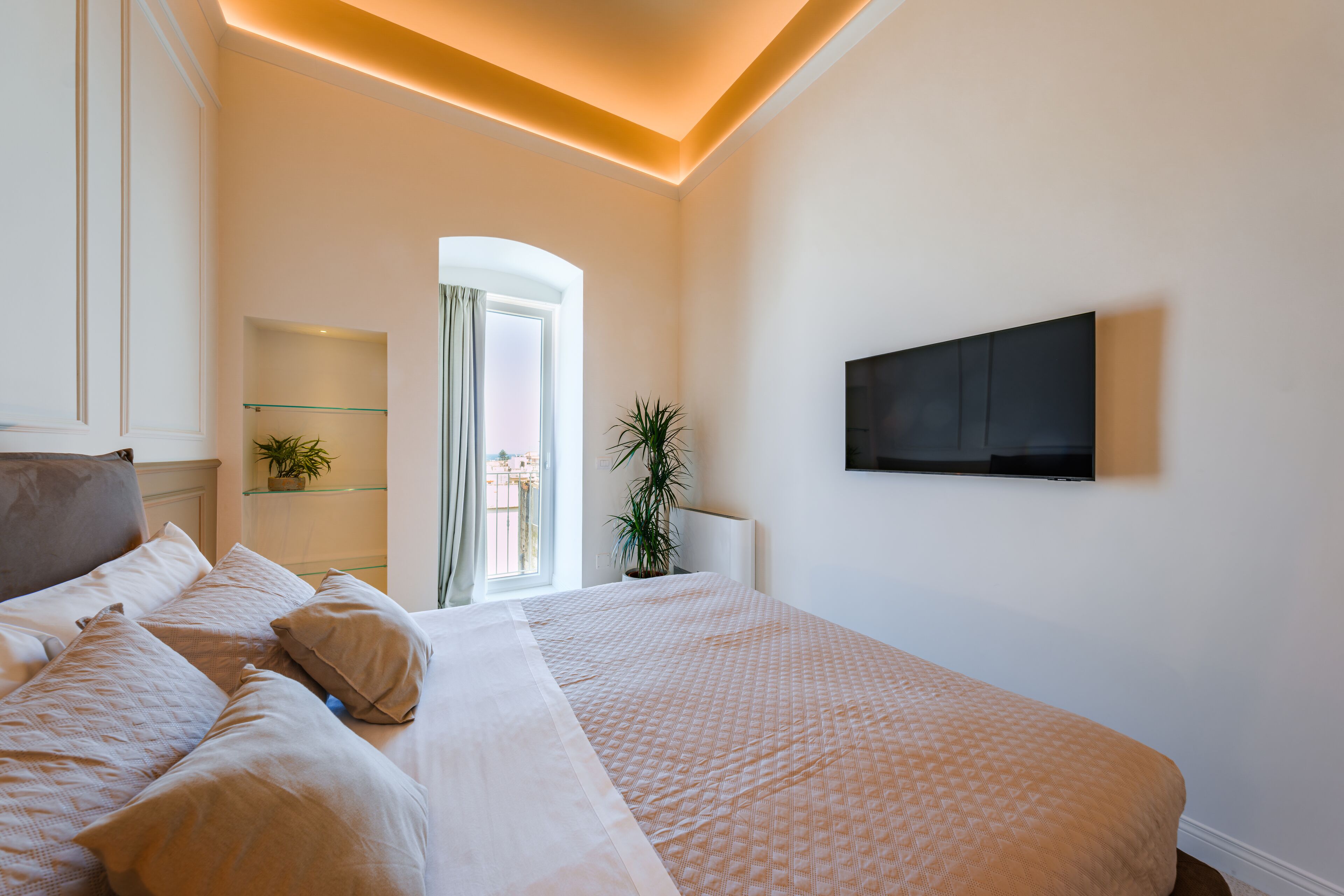 A hotel room with a large bed, a TV mounted on the wall, a potted plant, and a view of the outdoors through a window.