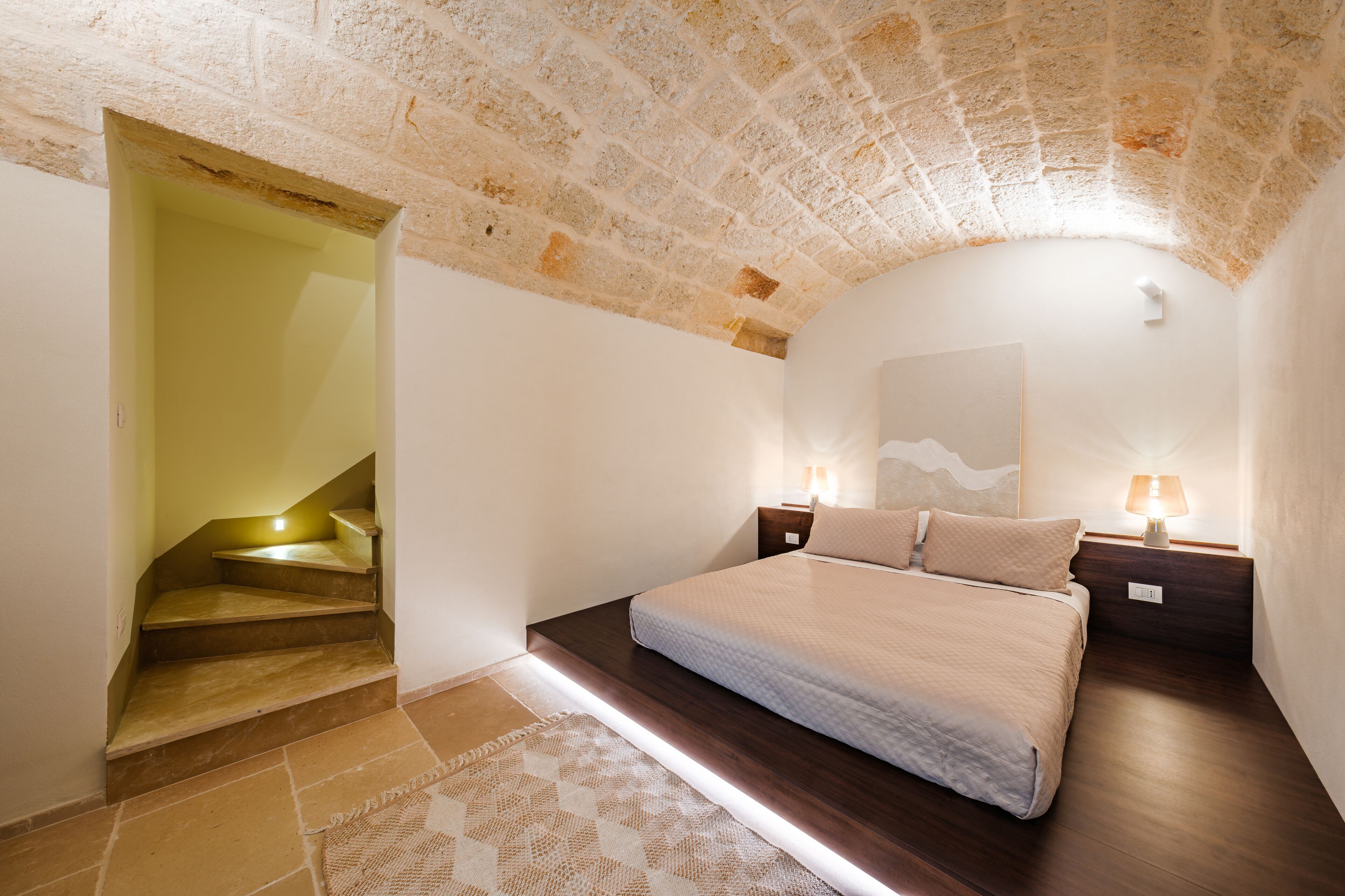 A bedroom with a bed, nightstands, a lamp, a staircase, and a stone ceiling.