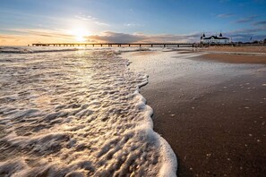 Ein Strand mit Wellen, einem Steg und einem Gebäude mit heller Fassade.