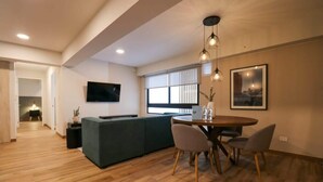 A modern living room with a dining area, wooden flooring, a sofa, a TV, and a window with blinds.