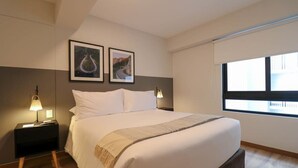 A bedroom with a bed, two bedside tables with lamps, a window with blinds, and two framed pictures on the wall.