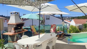 A poolside area with a barbecue grill, outdoor seating, umbrellas, and a view of the surrounding landscape.