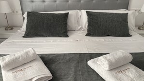 Una cama bien hecha con ropa de cama gris y blanca, dos almohadas grises y dos toallas blancas con texto.
