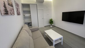 Una sala moderna con un sofá color beige, una mesa de centro blanca, un televisor de pantalla plana montado en la pared y una planta en maceta.