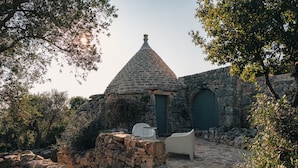 A stone building with a conical roof, a door, and a small table with a chair. - Masseria Minetta (Cisternino)