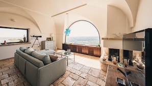 A spacious living area with a large window offering a view of the landscape, a cozy sofa, a wooden coffee table, and a fireplace with decorative items. - Masseria Minetta (Cisternino)