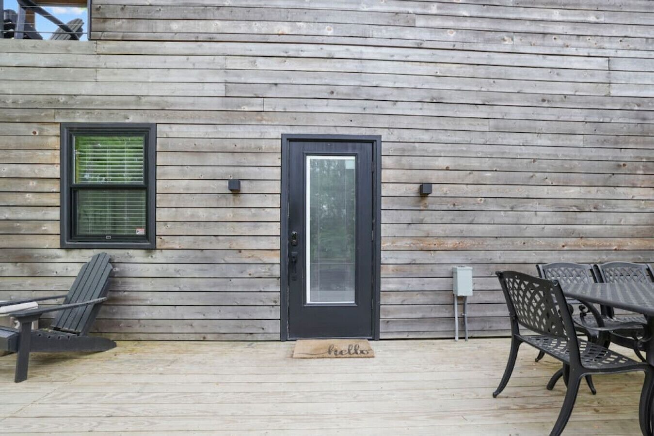A wooden exterior wall with a black-framed door, a window with blinds, and outdoor seating with metal chairs and a table.