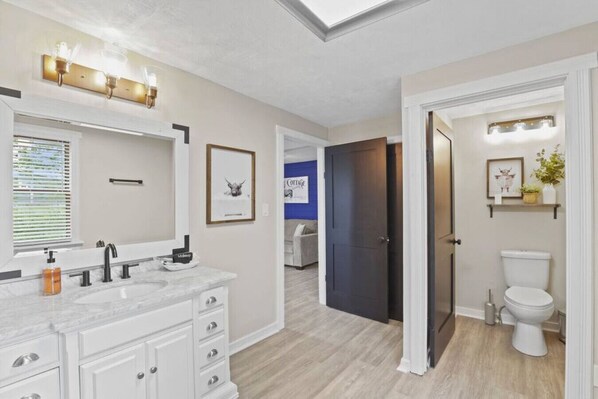 A modern bathroom with a large mirror, two sinks, and a toilet. There is a door leading to a bedroom with a bed and a blue wall. - Cattle Cottage: 2 minutes to the marina (Millersport)