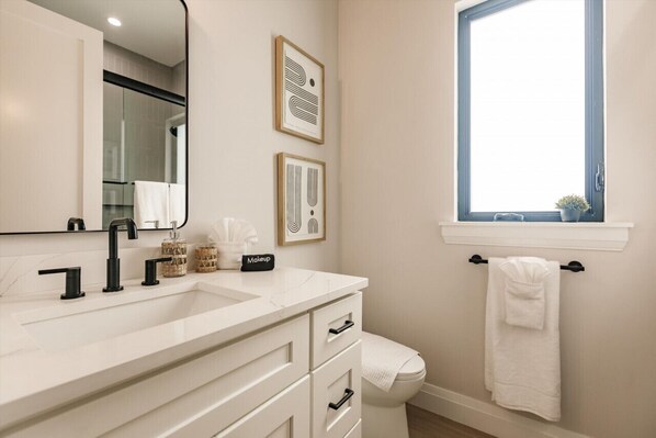 A modern bathroom with a white vanity, black faucets, and a large mirror. There are framed artworks on the wall and a window with a view.