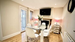 A modern dining area with a glass table and white chairs, a flat-screen TV mounted above, and a fireplace with pink lamps. - Mill House Townhouse (Newmarket)