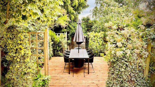 A wooden deck with a table and chairs, surrounded by lush greenery and climbing plants. - Mill House Townhouse (Newmarket)
