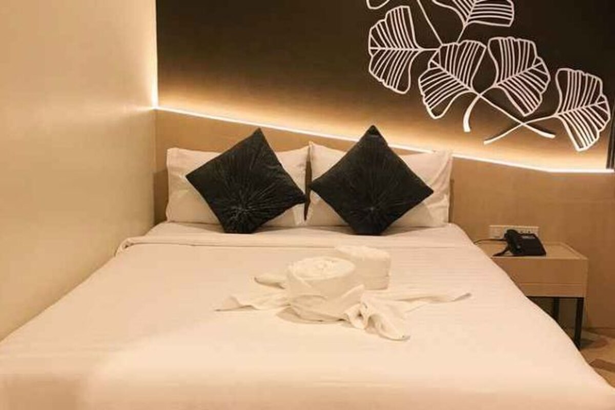 A hotel room with a neatly made bed, a bedside table with a phone, and a wall with a leaf-like light fixture.