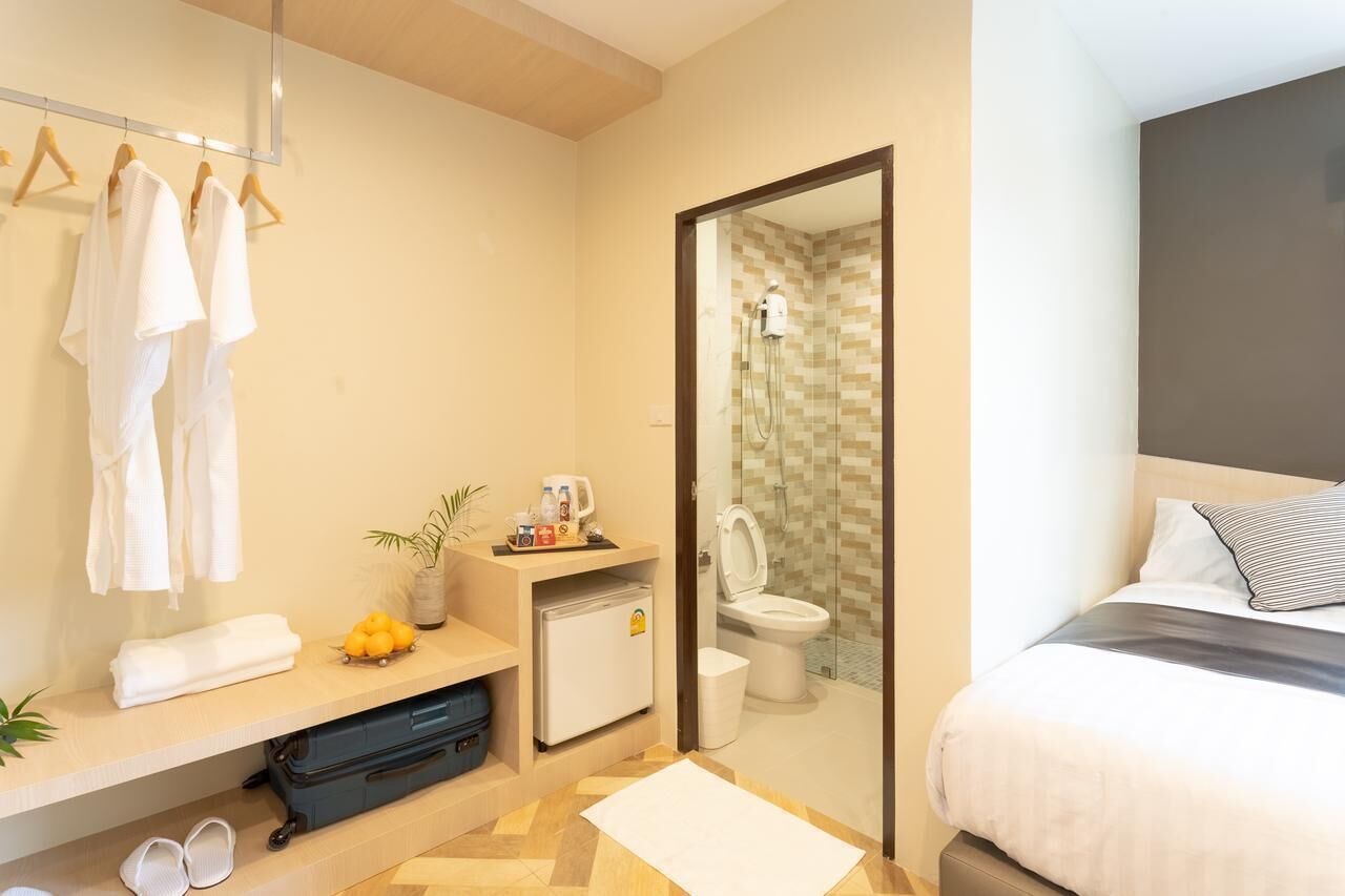 A modern hotel room with a bed, a small bathroom with a shower, a vanity with a mirror, and a shelf with towels and toiletries.