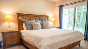 A neatly made bed with a wooden headboard, two bedside tables with lamps, and a view of greenery through a large window. - 3BR Okemo Escape: Premier Condo Steps to Trail (Ludlow)
