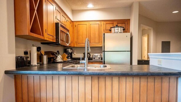 A kitchen with wooden cabinets, a stainless steel refrigerator, a microwave, and a sink. There are three stools in front of the counter. - 3BR Okemo Escape: Premier Condo Steps to Trail (Ludlow)