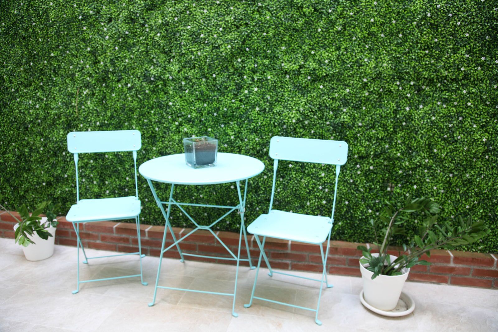 A small outdoor seating area with a round table and two chairs, set against a green hedge backdrop.