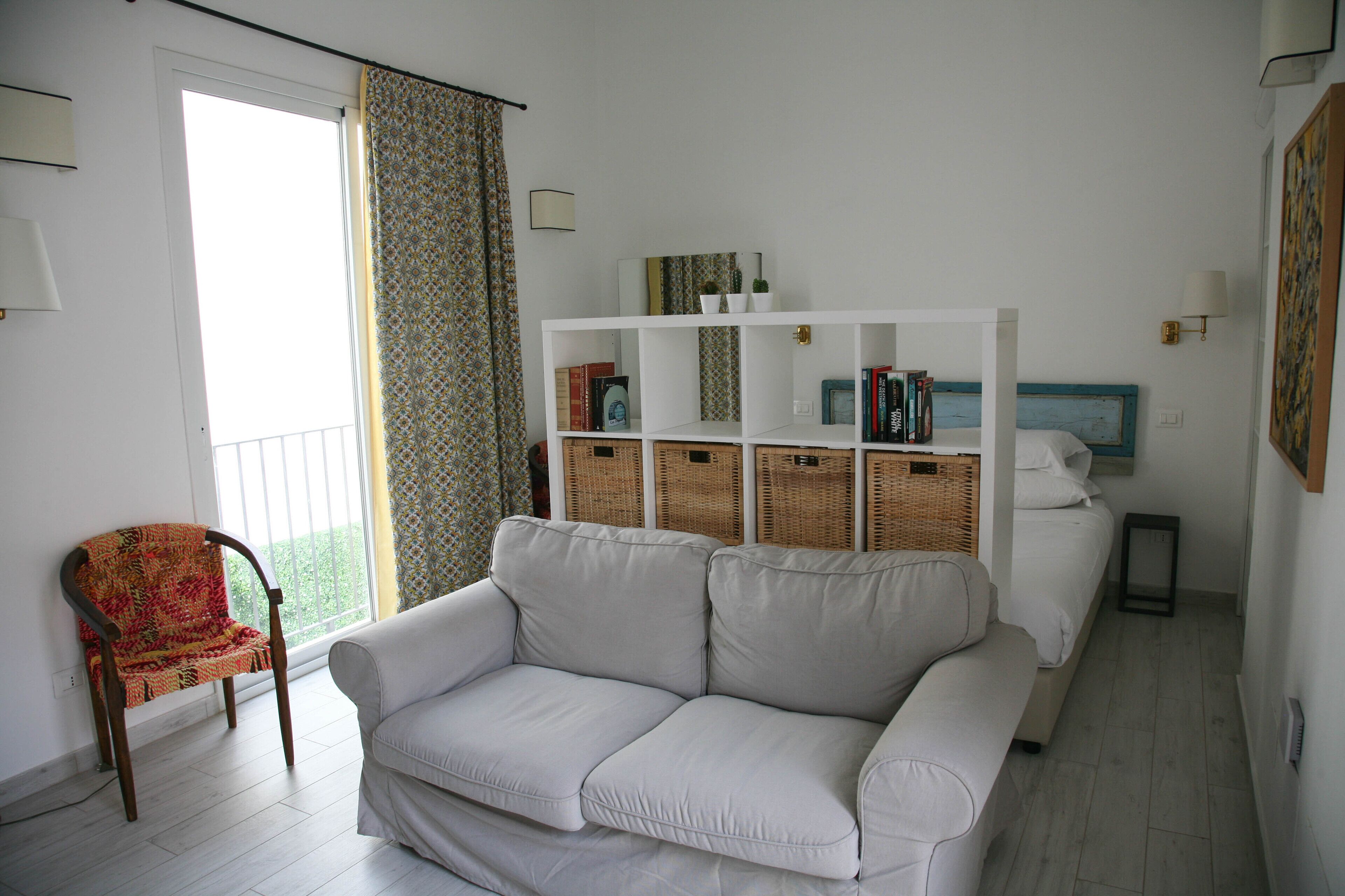 A room with a white sofa, a patterned armchair, a bookshelf, and a bed with a blue headboard.
