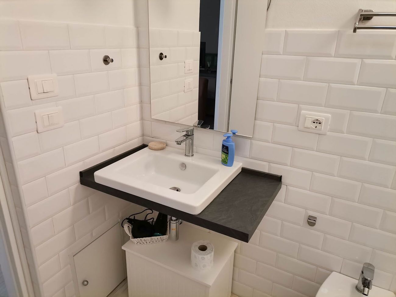 A bathroom with a white sink, a mirror, a black countertop, and a white tiled wall.
