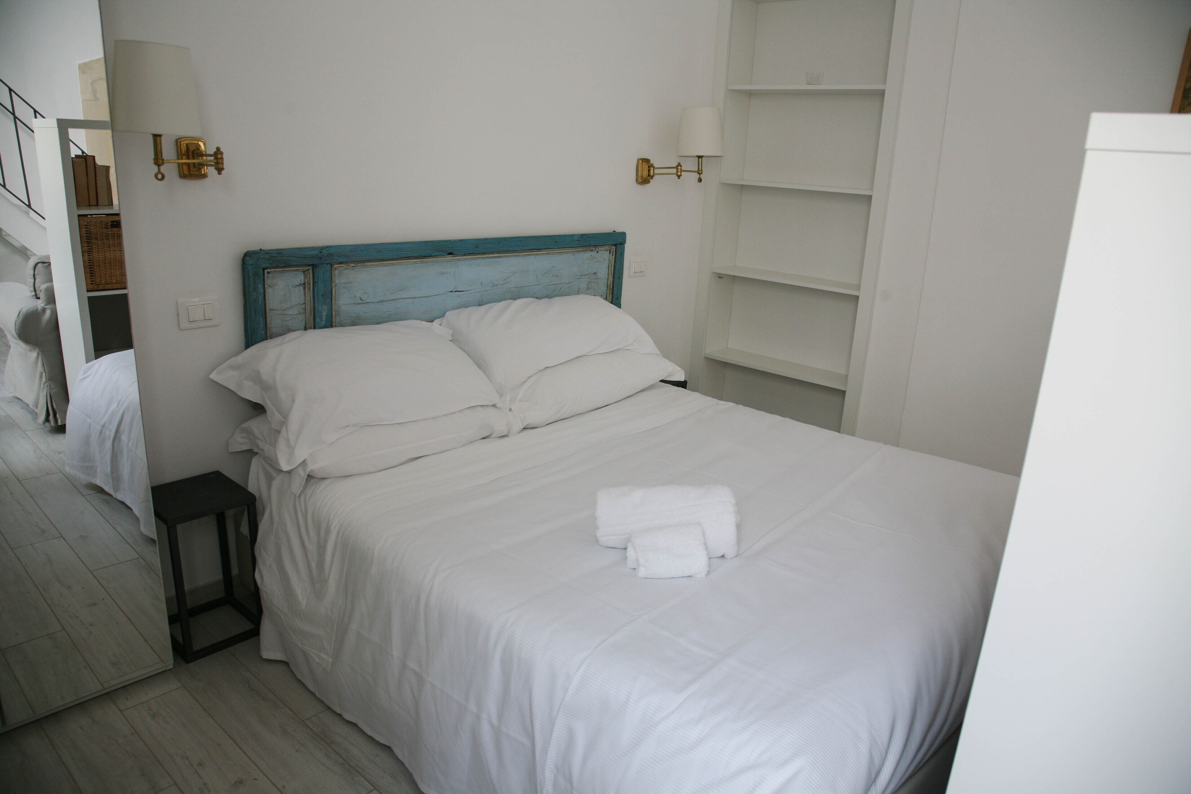 A neatly made bed with white linens and a blue headboard, a small bedside table, and wall-mounted lamps.