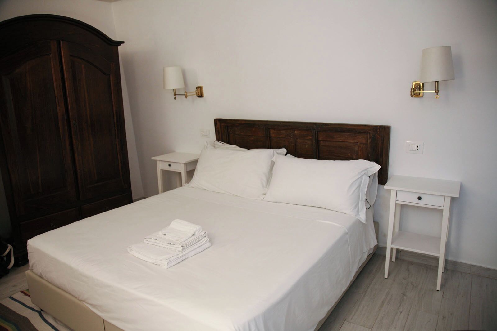 Una cama bien hecha con sábanas blancas y una toalla doblada, dos mesitas de noche con lámparas, y un armario de madera oscura.