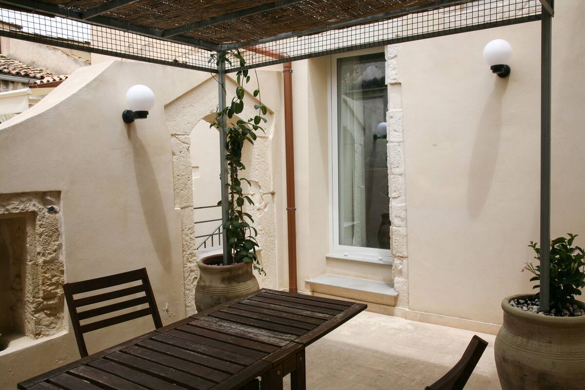 A covered patio with a wooden table and chairs, potted plants, and two wall-mounted lights.