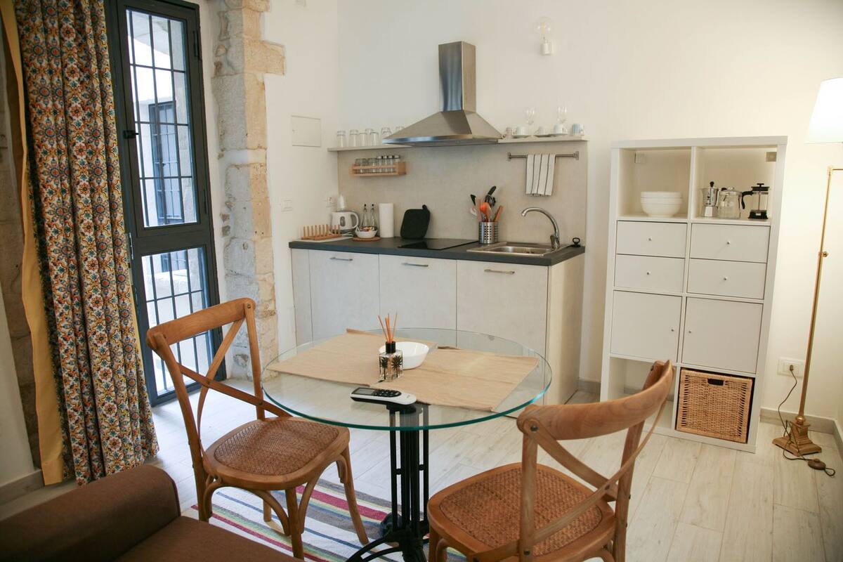 A compact kitchen with a round glass table, two wicker chairs, a built-in stove, and a small dining area.