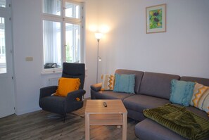Una sala de estar con un sillón gris, un sofá marrón y una mesita de madera. Hay una ventana con cortinas, una lámpara de pie y un cuadro en la pared.