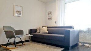 Een moderne woonkamer met een grijze bank, een schommelstoel, een klein wit tafeltje en twee ingelijste foto’s aan de muur.