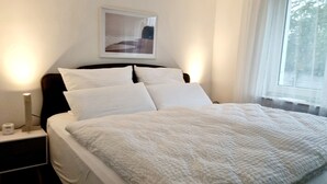 Een net opgemaakt bed met witte lakens en kussens, een nachtkastje met een lamp, en een ingelijste foto aan de muur.