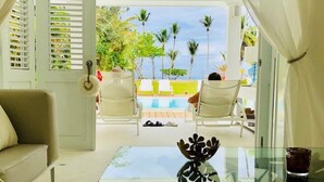 A room with a glass table, white curtains, a sofa, and a view of an outdoor pool area with chairs and palm trees.