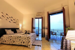 Un dormitorio con cama, mesita de noche, estante en la pared, balcón con barandilla y vista al mar.