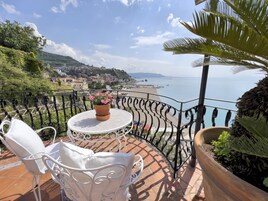 A balcony with a white metal table and chairs, a potted plant with pink flowers, and a view of the sea and a town in the background.